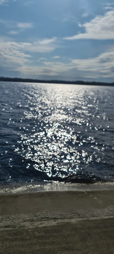 This image captures a vibrant waterscape scene, rotated 90 degrees clockwise. On the left, a bright blue sky is adorned with soft, white cumulus clouds, suggesting fair weather. The central and largest portion of the image is dominated by a body of water, likely a river or lake, such as the Daugava River in Ikšķile, Latvia. The water's surface is alight with numerous sparkling reflections of sunlight, creating a dynamic pattern of bright, out-of-focus bokeh circles across its dark blue expanse. A faint, dark strip near the horizon separates the water from the sky, possibly indicating a distant shoreline or treeline. To the far right, a textured, weathered concrete or stone structure, possibly part of a pier, embankment, or wall, runs vertically along the edge of the water. The strong glare on the water indicates bright sunshine, suggesting a clear day during morning or afternoon hours. No specific activities, individuals, or visible text are present in the static scene, which conveys a sense of serene natural beauty.