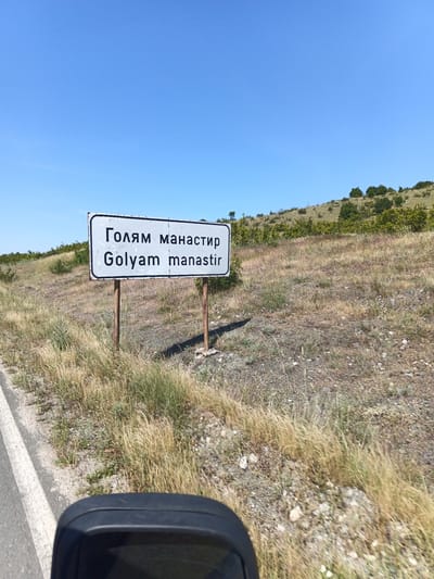 A sign on two metal posts reads "Голям Манастир" and "Golyam manastir" in white text with a black border. The sign is located in a rural area with dry grass and bushes. A road is visible on the left side of the image. The sky is clear and blue. The scene appears to be in Golyam Manastir, Bulgaria.