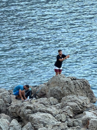The image shows two men fishing on a rocky coastline. The man on the left is crouched down on the rocks, facing away from the camera, and appears to be preparing fishing bait. He is wearing a blue t-shirt and blue shorts. Next to him is a black fishing tackle bag and a fishing rod. The man on the right is standing on a higher rock, facing the water, and is actively fishing with a rod. He is wearing a black t-shirt and red and white striped shorts. The sea behind them is choppy and appears to be a deep blue color. The rocks are rough and gray. The scene is likely located in Baunei, Italy, known for its rugged coastline and clear waters.