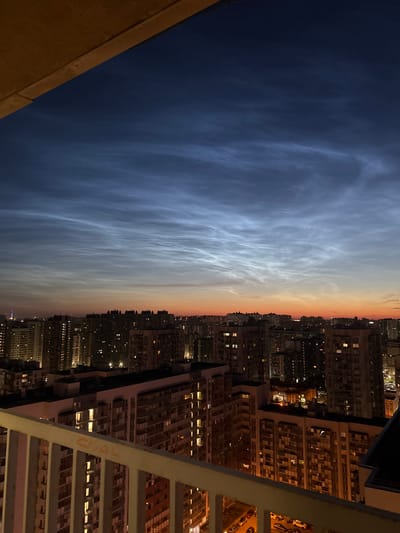 The image depicts a city skyline at dusk, likely in Oreshek-3, Russia, given the context. The sky is dominated by noctilucent clouds, which appear as wispy, luminous formations against a gradient of deep blue and warm orange hues. Below the sky, a dense arrangement of multi-story apartment buildings fills the midground, their windows illuminated with warm, yellow light, indicating occupancy. The foreground features a portion of a balcony railing, painted white and showing some signs of graffiti, with the letters "CRAL" visible in orange. Cars are parked in an outdoor lot visible in the lower right of the image. The overall impression is one of urban life unfolding under an extraordinary atmospheric phenomenon.