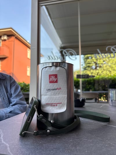 The image depicts a table at what appears to be an outdoor cafe, with a reflective window showing the exterior. A metal napkin holder containing Illy-branded napkins is on the table. The napkin holder is positioned near a green phone case attached to a lanyard. The background visible through the window shows a building with an orange exterior and green trees. Based on the Illy branding and the buildings, the location is Longone al Segrino, Italy. A person is partially visible, wearing a denim jacket.