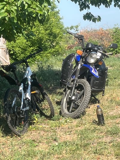 Two vehicles, a white bicycle and a blue and black motorcycle, are parked on grassy ground in an outdoor setting. The motorcycle has a blue fuel tank with white and red decals, black saddlebags, a round headlight, and a black handlebar with orange turn signals. A bottle filled with dark liquid stands upright on the ground near the motorcycle's front wheel. The bicycle is positioned to the left of the motorcycle, and it is white with black accents, including fenders and a chain guard. A black bag is attached to the bicycle's frame. Lush green trees and vegetation surround the vehicles, with a clear blue sky visible above. The scene appears to be in a rural or semi-rural area.