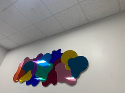 This media file depicts a wall art installation on a white wall. The art is composed of irregularly shaped, overlapping pieces with a mirrored, iridescent surface that reflects the surrounding environment and the ceiling lights. The colors visible include pink, orange, yellow, green, teal, blue, purple, and maroon. Some pieces are brighter and more reflective than others, suggesting variations in material or lighting. The ceiling above is a standard drop ceiling with square tiles and recessed lighting fixtures, typical of an office or commercial space. The art is arranged in a horizontal, somewhat clustered formation across the wall. There are no people or animals visible in the image, and no specific activity or event is taking place. The lighting suggests it is daytime, but the indoor setting provides no information about the weather. The location context is Chesapeake, United States of America, but there are no visual cues in the image to confirm this. No text is visible in the media file.
