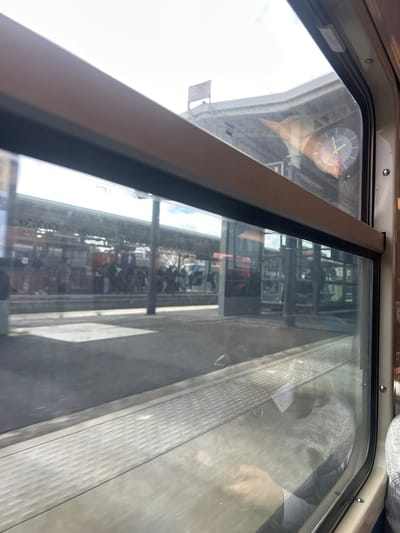 The image depicts a view from inside a train, looking out through a window at a train station platform in Bondy, France.

**Scene Description:**
The perspective is from a passenger seat, with the beige frame of the train window dominating the foreground and sides. The window itself shows reflections from the train's interior, including the hands and upper torso of a person, possibly the photographer, wearing a light-colored sweater over a striped shirt in the lower right quadrant.

Outside the window, a train station platform is visible. A group of approximately twenty to thirty individuals are standing on the platform, primarily grouped in the mid-ground, facing generally towards the right side of the frame. The platform features tactile paving near its edge. Overhead, a large, metal-framed canopy structure extends over the platform, supported by vertical pillars. Above and to the right of the platform, a large, circular analog clock with yellow and black hands is mounted on an external building or support structure, indicating a time close to 1:55. The sky, showing light cloud cover, is visible above the station's architecture. Further in the background, beyond the platform, additional structures and possibly another set of tracks or train are faintly discernible. The scene is illuminated by daylight, casting some shadows on the platform.