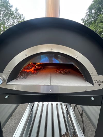 An outdoor "Vulcan+" pizza oven, predominantly black with stainless steel accents and a copper chimney, is positioned outdoors in Hornsea, United Kingdom. Inside the oven, a wood fire burns on the left, casting an orange glow, while a single cooked pizza rests on the light-colored refractory tiles on the right. A temperature gauge is visible on the lower-left front of the oven. A reflection of an individual, likely the photographer, wearing a light-colored shirt, is discernible in the oven's interior glass or reflective surface. The oven is supported by a black frame with a silver slatted shelf beneath. The immediate ground appears to be paved, with a partial view of a metal folding table leg on the far right. The background shows mature trees under an overcast sky.