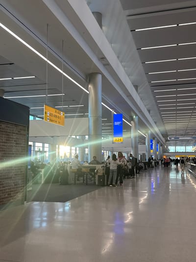 An airport hallway with a high ceiling and modern lighting. People are seated and standing, likely waiting at gates. Directional signs indicate gate numbers and baggage claim. The scene is well-lit and appears to be in a busy terminal. A brick wall adds texture to the otherwise modern interior. The orientation of the image is vertical.