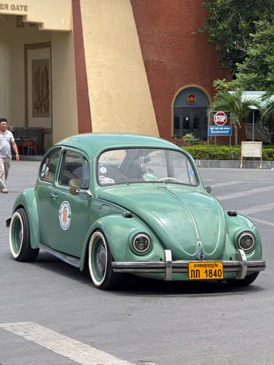 In Lao Bảo Commune, Vietnam, a vintage green Volkswagen Beetle is parked on a paved road. The car features white-walled tires, chrome bumpers, and a yellow license plate with Lao script and the number "1840". A man in a light-colored polo shirt and pants walks away from the car in the background. Behind the car, a building with a tan and red brick facade is visible. A "STOP" sign is posted near the building, along with some greenery and palm trees. The overall setting appears to be an outdoor area, possibly a commercial or administrative district, given the presence of the building and signs.