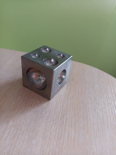 A silver, cubic metal object with hemispherical indentations on its surfaces sits on a light-colored wooden table. The object has indentations on the top and visible sides. The background is a plain green wall. The image is taken from a slightly elevated angle, looking down at the object. The lighting suggests an indoor environment. There are no people present, and no activity is taking place. The location is not specified as Vilyuysk, Russia.