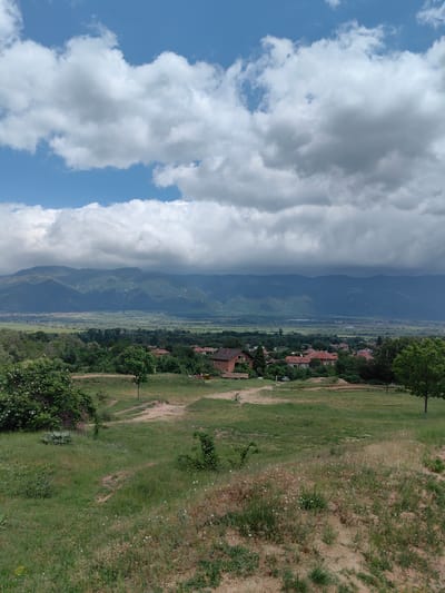 The image shows a landscape in Slatina, Bulgaria. In the foreground, there is a grassy and sandy area, dotted with small white flowers and sparse vegetation. In the midground, a small village is nestled amongst trees, with houses visible, characterized by red tile roofs. In the background, a wide valley stretches out towards a mountain range under a sky filled with heavy, layered clouds. The light is diffused, suggesting overcast conditions.