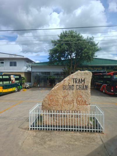 This image depicts an outdoor scene at what appears to be a bus station or transportation hub in Tân Phú 2, Vietnam. The dominant subject is a large, rough-hewn stone monument with text carved into it. The stone is surrounded by a white metal fence, suggesting it's a significant landmark or memorial.
The text on the stone monument is in Vietnamese. The top line reads "TRẠM" and the second line reads "DUNG CHÂN". Below this, in a more decorative script, are other phrases, including "Trạm Dừng Chân" and possibly names or additional information.
In the background, a modern building with a green roof and glass-fronted windows is visible. A bright red and black bus is parked in front of this building, suggesting it's operational. Further to the left, a lighter colored building with a white exterior and windows is seen.
To the right of the stone monument, a yellow and green bus is parked, its doors open, with people potentially inside or alighting. The ground is paved with concrete, marked with yellow lines, typical of a parking lot or road. A mature tree with lush green foliage stands near the stone monument and the building.
The sky above is partly cloudy with patches of blue, indicating it's daytime. The overall impression is a functional, perhaps slightly rural, transportation facility. There are no distinct emotions conveyed, but the scene appears to be active with the presence of buses and a building that suggests services. The lighting suggests it is midday.