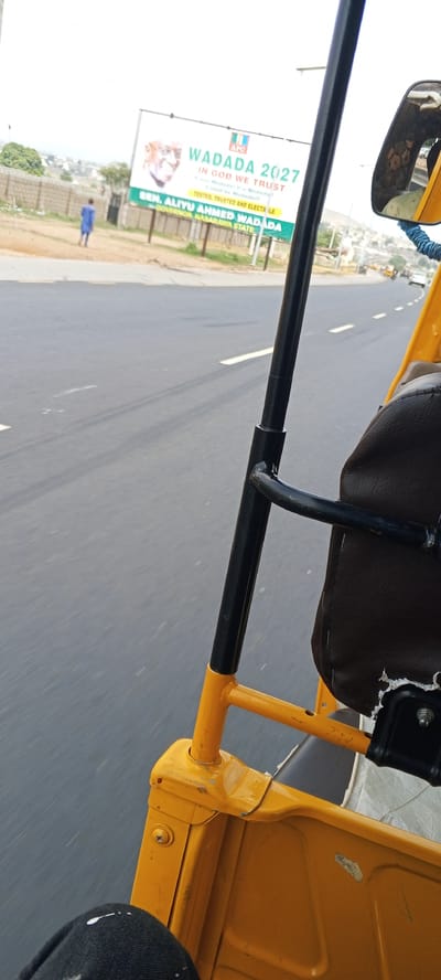 The image shows a view from inside a yellow auto-rickshaw, commonly known as a "Keke" or "Tricycle", moving along an asphalt road. The road has white lane markings. On the roadside, there is a billboard promoting "WADADA 2027" with the text "IN GOD WE TRUST", and "SEN. ALIYU AHMED WADADA GOVERNOR NASARAWA STATE". A person is walking near the billboard. The location is Uke, Nigeria.