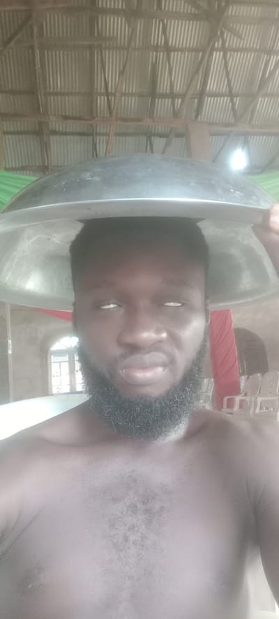 A shirtless man with a dark beard and white eyes holds a large metal bowl on his head. He is indoors, possibly in a simple structure with a corrugated metal roof supported by wooden beams. A window and several chairs are visible in the background. The location is Bende, Nigeria.