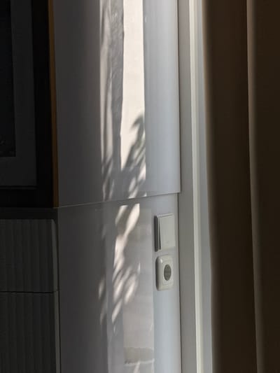 The image shows a wall with two electrical outlets and a light switch. The wall is white and has shadows cast on it from what appears to be plants. To the left of the wall is a dark, textured surface, possibly furniture or a cabinet. To the right, there is a brown curtain. The lighting suggests it is daytime, with sunlight coming from the right. The scene appears to be indoors. The location of Tanjungmeru, Indonesia is not identifiable from the visual information present in the image.