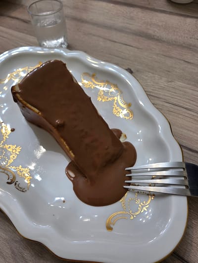 A slice of chocolate cake, covered in melted chocolate, sits on a white, ornate plate. A fork rests on the right side of the plate, partially touching the cake. In the background, a small glass of clear liquid is visible. The scene is set on a wooden table. The image was taken in Yalangach, Uzbekistan.
