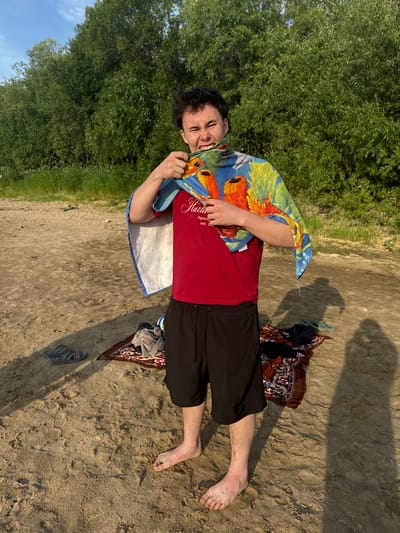 A young man with dark, messy hair stands on a sandy beach, facing the camera. He is wearing a red t-shirt and black shorts. A colorful beach towel with a parrot design is draped over his shoulders and wrapped around his neck, and he is using it to playfully tug on his lower lip. His facial expression is one of playful discomfort. His feet are bare and dusty. In the background, a dense line of green trees separates the beach from the sky. Scattered on the sand are a pair of sandals, a folded patterned rug, and some clothing. The lighting suggests it is late afternoon or early evening, casting long shadows on the sand. The scene appears to be a casual, outdoor setting, possibly during a warm weather outing near a body of water. The location is identified as Рябинино, Russia.