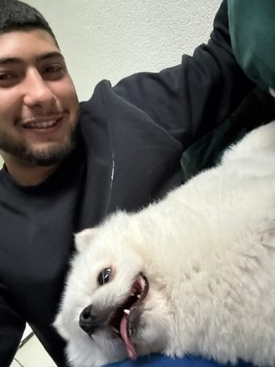A close-up, waist-up, selfie-style photograph shows a young adult male, appearing to be of Middle Eastern descent, with dark hair and a beard, smiling broadly as he holds a small white dog. The male is wearing a black hooded sweatshirt. The dog, possibly a Pomeranian or similar breed, is white and fluffy, with its mouth open and tongue lolling out, looking towards the camera. The background is a plain, textured wall, likely indoors, and a dark green blanket or clothing item is visible on the right side of the frame. The lighting is bright and direct, suggesting artificial indoor lighting. The overall scene conveys a casual and affectionate interaction between the man and his pet. The location is specified as Lausen, Switzerland.