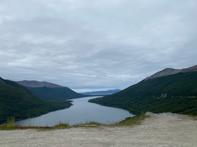 A wide river, its waters reflecting the cloudy sky, flows between wooded hills. The slopes are densely vegetated in dark green, contrasted by patches of bare rock on their upper reaches. A slightly elevated, sandy foreground hints at the viewing point. The atmosphere is muted, dominated by overcast skies and a somber color palette. Distant mountains can be seen where the river fades into the horizon.