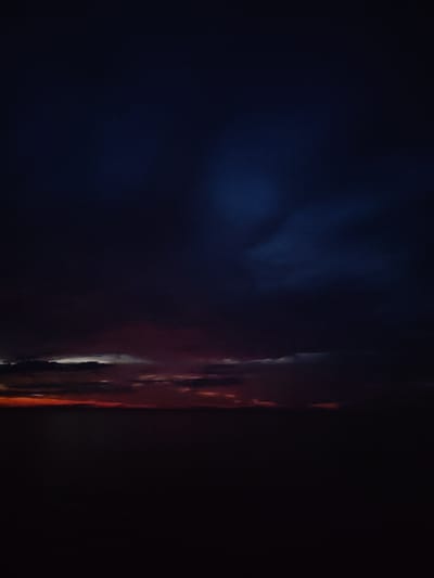The image depicts a dark, ominous sky over a body of water at dusk or dawn. The sky is filled with heavy, bruised-looking clouds, tinged with deep blues and purples, transitioning to a band of reddish-orange at the horizon. The water below is almost entirely black, reflecting very little of the fading light. There are no visible signs of human activity, structures, or distinct geographical features of the city of Kvartsitnyi, Russia. The scene is dominated by the moody atmosphere of the sky and the obscured landscape.