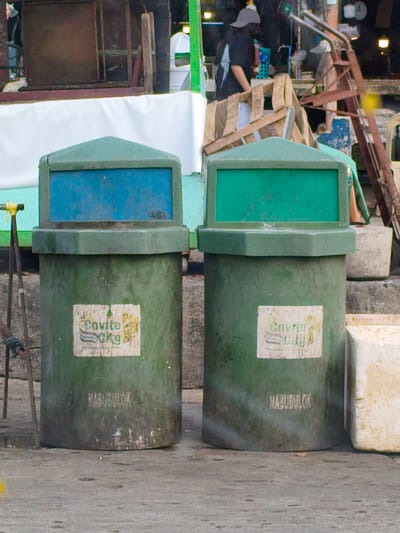 Two green trash bins with blue and green lids stand on a paved surface, possibly outdoors. The bin on the left has a blue lid and a rectangular label with text and imagery. The bin on the right has a green lid and a similar label. Below the labels, the word "NABUBULOK" is visible on both bins, which translates to "decomposing" or "rotting" in Tagalog, indicating they are for organic waste. A white tarp or fabric is stretched behind the bins. In the background, there are wooden structures, a person wearing a dark shirt and a cap, a metal cart, and other elements of what appears to be a marketplace or a busy outdoor area. The lighting suggests it could be daytime, possibly overcast or evening. The overall scene is one of a public space with waste management facilities. The labels on the bins read "Cavite City", providing a clear location cue.