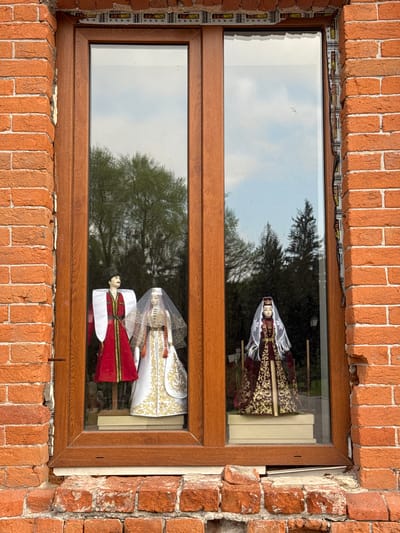The image shows a window framed by brickwork, likely located in Akhieli, Georgia, given the presence of traditional Georgian dolls displayed inside. The window is a divided pane style with a brown wooden frame. Inside, on a shelf, are three dolls dressed in traditional Georgian attire. The top doll wears a dark, patterned robe with a white lace headdress. The lower dolls include a figure in a white and gold robe with a matching headdress and a figure in a red and white robe. The background visible through the window is a blurred reflection of trees and sky, suggesting an outdoor setting. The brickwork surrounding the window appears aged, adding to the setting’s character.