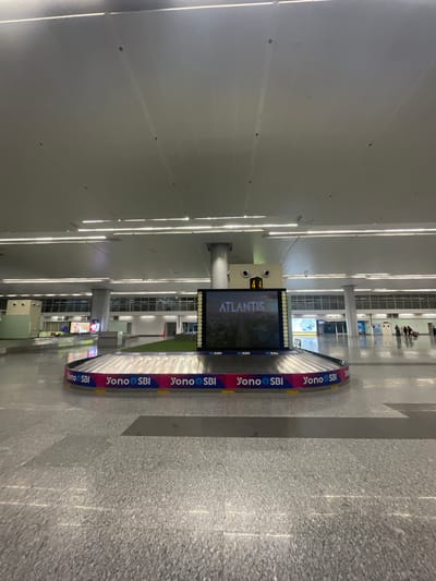 The image depicts an interior view of an airport baggage claim area, specifically at Swami Vivekananda Airport (R new) in Raipur Khurd, India. In the foreground, a baggage carousel is prominently featured, adorned with "Yono SBI" branding. Above the carousel, a large digital display screen shows "ATLANTIS" in large white text, with a smaller number "44" displayed above it. The floor is a polished, speckled gray material. In the background, through large windows, a seemingly empty airport terminal can be seen. A few individuals are visible in the far background, walking. The overall environment is clean, well-lit with fluorescent lighting from the ceiling, and appears to be at a low traffic time.