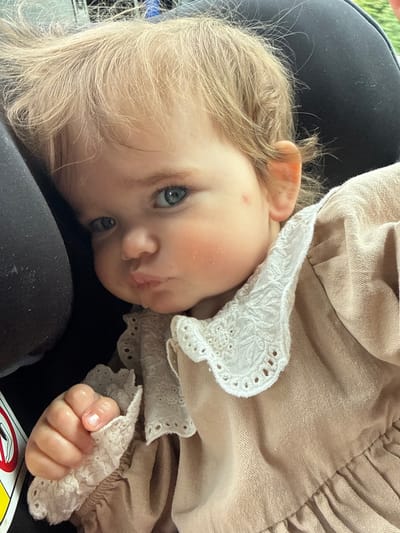 A close-up, eye-level shot shows a baby with blond hair and blue eyes. The baby is wearing a tan dress with white lace trim. The baby's left hand is visible, holding onto the lace. The baby is in a car seat, indicated by the black headrest and a portion of a warning label visible on the left. The lighting suggests it is daytime, possibly outdoors. The image is taken in Limburg an der Lahn, Germany.
