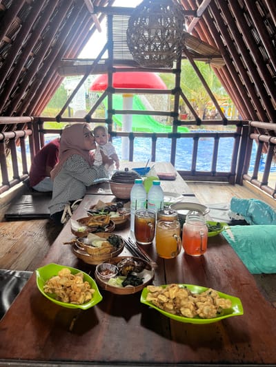 The image depicts an indoor-outdoor dining area, likely a traditional open-air gazebo or hut, within a water park or resort setting in Juwana, Indonesia. The structure is constructed with prominent wooden or bamboo beams forming an A-frame roof and open sides, offering a view of a swimming pool.
In the mid-ground, a woman wearing a light brown headscarf (hijab) and a black and white checkered shirt is seated, holding and kissing an infant. The infant is dressed in a striped top. Another individual, largely obscured, is seated to the woman's left.
The foreground is dominated by a long, dark wooden table laden with food and beverages. Multiple dishes, served in woven bamboo or wooden bowls lined with paper, contain various fried items, possibly poultry or seafood, accompanied by vegetables and dipping sauces. Two bright green, boat-shaped plates hold piles of golden-brown fried snacks. Several clear glasses contain beverages of varying colors: dark brown (likely iced tea), orange, and reddish-pink. Two plastic bottles of water are also visible, along with a dark, lidded serving dish.
Beyond the dining area, a large blue swimming pool is visible, featuring recreational water park elements. These include a prominent red mushroom-shaped structure with water falling from it, and a green water slide. Distant foliage, possibly palm trees, and other pool structures are also discernible under bright outdoor lighting.