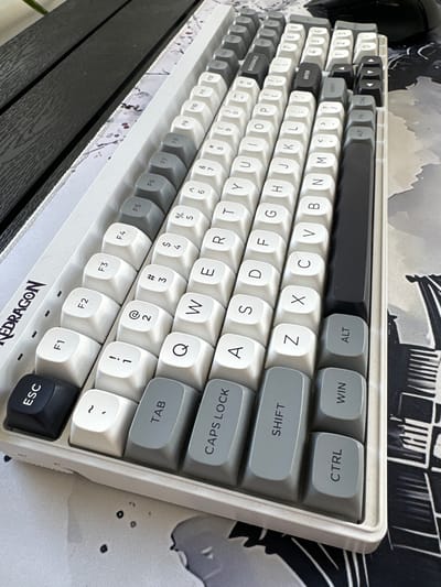 The image shows a close-up of a white mechanical keyboard with a two-tone keycap design, featuring white and grey keys. The keyboard is positioned on a desk with a dark wooden surface to the left and a patterned desk mat with grey and white abstract designs underneath the right side. The Redragon logo is visible on the top left edge of the keyboard. The keyboard is angled, with the top right corner closer to the viewer. Function keys (F1-F12) are arranged in the top row, followed by number keys, and then the alphanumeric keys. Modifier keys such as ESC, TAB, CAPS LOCK, SHIFT, CTRL, WIN, and ALT are visible on the left and bottom rows. The keys are illuminated by ambient light, casting subtle shadows. No people are present in the image. The setting appears to be an indoor workspace. The specific location of Moortown, Ireland, is not evident from the image itself.