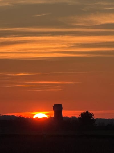 The image displays a sunset over a rural landscape in Broadland, Norfolk, United Kingdom. The sky is characterized by horizontal bands of vibrant orange and red hues near the horizon, transitioning to lighter orange and muted grey-orange at higher altitudes, indicative of layered clouds. A large, bright, semicircular sun is prominently positioned centrally on the horizon, its lower portion obscured by the dark, silhouetted foreground. Silhouetted against the illuminated sky is Strumpshaw Mill, a distinctive, dark-toned tower mill structure, appearing truncated without a cap. To the immediate right of the mill, a cluster of dark, silhouetted trees is visible. The foreground consists of a dark, indistinct horizontal plane, suggesting an open field or terrain. No people or discernible activities are present in the scene.