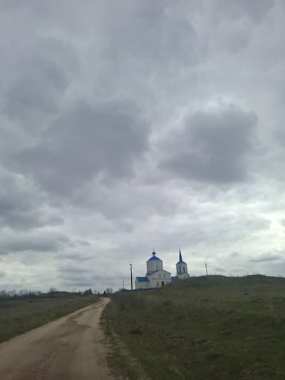 A landscape photograph captures a quiet, rural scene in what appears to be Yablonovo, Russia, under an overcast sky. The image is dominated by heavy, grey clouds, suggesting a cloudy day, possibly with cool weather.

In the midground, a white church-like building with distinctive blue onion domes and spires stands out against the muted backdrop. The church features multiple sections and visible windows. Utility poles with wires are visible near the building, extending alongside a dirt road.

The dirt road meanders from the foreground on the right side of the frame, curving towards the center-left. To the right of the road, a gently sloping hill covered in dry, sparse grass rises, suggesting either late autumn or early spring. Distant, bare deciduous trees are faintly visible on the horizon line.

No people or specific activities are visible in the frame, contributing to a sense of solitude. The architectural style of the church, with its white walls and blue onion domes, is characteristic of Russian Orthodox churches, aligning with the provided location context. The overall mood is one of stillness and quiet reflection under a somber sky. There is no visible text.