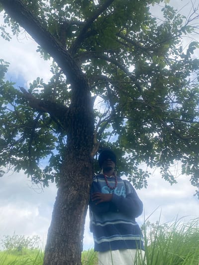 A person stands under a large tree in Unwana, Nigeria. The person is wearing a blue and white striped sweater over a white shirt, and a black head covering. Red beads are around their neck. The tree has a rough, dark brown trunk and branches spread out, covered in green leaves. The background shows a cloudy sky and lush green grass.