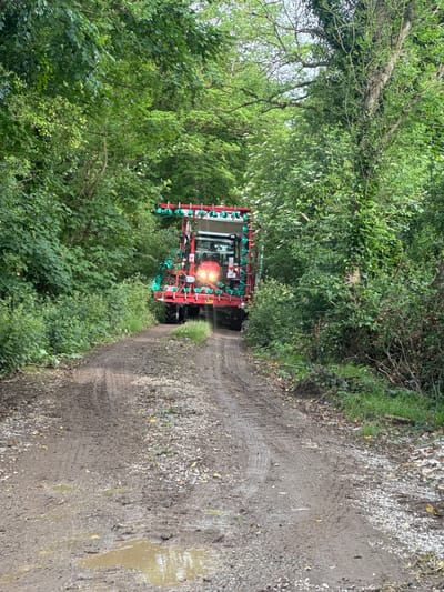 An agricultural tractor, painted red with its rear lights illuminated, is the primary subject of this image. The tractor is equipped with a green-nozzled sprayer boom, which is partially extended or folded, visible across its rear. The vehicle is situated on a muddy, unpaved track or lane, which shows distinct tire tracks and patches of gravel. Several puddles of standing water are present in the muddy sections of the track, indicating recent wet weather.
Dense, lush green foliage, consisting of trees and bushes, lines both sides of the track, creating a verdant, almost tunnel-like effect. The overall setting is rural and natural. The lighting suggests it is daytime, possibly overcast, given the lack of strong shadows, though the tractor's lights are on. There are no people visible. The specific location provided is Cross Keys, Ireland. No discernible text is visible on the vehicle or in the surroundings.