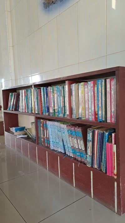 The image shows a low-angle, full shot of a dark reddish-brown wooden bookshelf filled with books. The bookshelf is positioned against a wall covered in cream-colored square tiles. A partial view of a mural or decorative pattern is visible at the top of the tiled wall. The bookshelf has two main shelves stacked with books. The top shelf is filled with books of various sizes and colors, predominantly with intricate patterns and Arabic script on their spines. Some of the books are leaning slightly. The bottom shelf is also filled with books, many of which are smaller and appear to be part of a collection, featuring similar decorative spines with Arabic text. In the lower left of the bottom shelf, a stack of smaller, lighter-colored books or pamphlets is visible, some of which are blue. The floor in front of the bookshelf is tiled with light gray square tiles. The lighting suggests an indoor environment, possibly a library or religious institution. There are no people visible in the image, and no actions or interactions are depicted. The overall impression is one of organized storage of books. The image does not contain any specific visual cues that would indicate the location in Cotbadatunong, Indonesia.