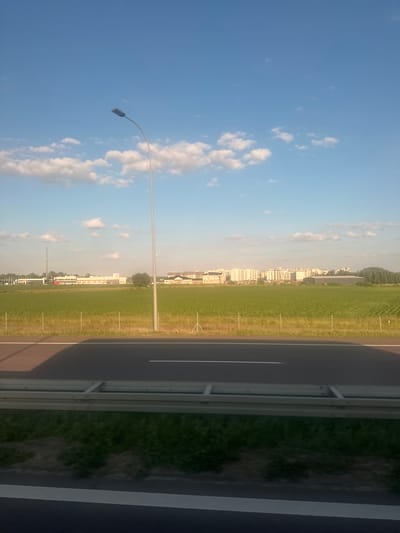This is a vertical, eye-level, outdoor, daytime photograph of a landscape with a road in the foreground and buildings in the distance. The sky is a clear blue with scattered white clouds. A tall, grey lamppost stands prominently in the left-center of the frame.
The middle ground features a vibrant green field of crops, possibly corn, with a row of thin, vertical poles running horizontally across it. Beyond the field, a cluster of buildings forms a distant skyline. The buildings are predominantly light-colored, appearing to be residential or office structures with multiple windows. To the far left, a tall communications tower is visible, and further to the right, there are some trees.
The foreground is dominated by a dark grey asphalt road with a white line marking its edge. A silver guardrail separates the road from the grassy verge. The scene appears to be located on the outskirts of a city, possibly Garwolin, Poland, given the architectural style and agricultural landscape. There are no people or animals visible in the frame, and no actions or interactions are depicted. The overall atmosphere is serene and quiet.