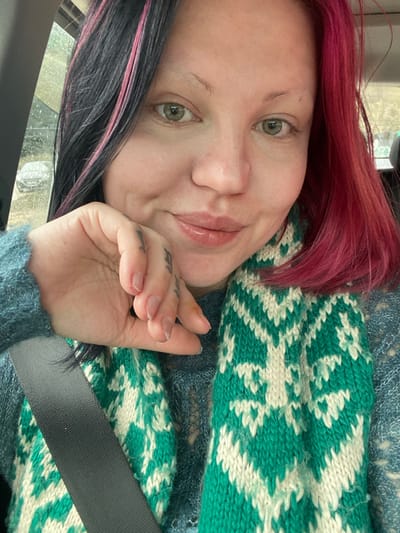 A close-up portrait depicts an individual, appearing female, inside a vehicle, located in Kruče - Kërruçi, Montenegro. The person has light skin, light green eyes, and a slight smile with glossy lips. Hair on the viewer's left is dark with pink streaks, while hair on the viewer's right is vibrant magenta. The individual wears a dark teal or blue knitted sweater with an open-weave pattern. A knitted scarf, featuring a geometric pattern in teal/green and off-white, is wrapped around the neck. A black seatbelt strap is visible across the individual's chest. Tattoos are present on the fingers of the left hand, which is positioned with fingers near the chin area. The background, blurred and seen through a vehicle window, suggests an outdoor scene, possibly overcast, with a very faint outline of what appears to be a parked car, potentially covered in snow or frost. The individual is looking directly at the camera.