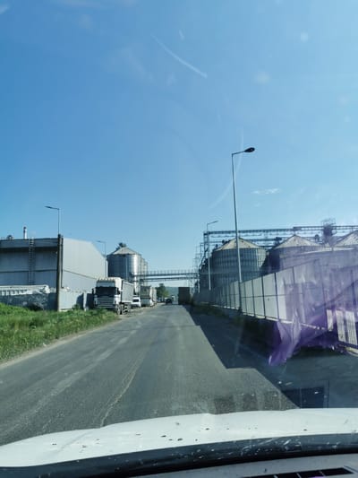 The image shows a straight road lined with industrial structures and parked trucks, possibly located in or near Razdelna, Bulgaria, given the presence of grain silos and associated facilities. The sky is clear and blue. There are several metal silos on either side of the road connected by overhead walkways. A row of semi-trailer trucks is parked on the left side of the road next to a large metal building with a ladder. Street lamps line the road. The view is taken from inside a vehicle, evident from the dashboard and windshield obstructing the lower portion of the frame. There are no people visible in the frame. The overall environment appears industrial, likely a storage or processing area for agricultural products.