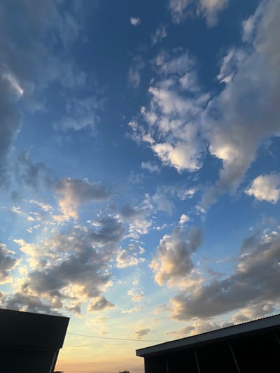 The image shows a partially cloudy sky over a village. The sky is a gradient of blue with white and gray clouds. The sun is setting, casting a warm glow on the clouds. In the foreground, there are silhouettes of two buildings, possibly houses, with dark roofs and walls. A thin wire stretches across the sky between the buildings. The location is identified as Ленинское сельское поселение, Ukraine. There are no people or animals visible in the image.