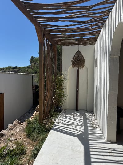 This outdoor image, captured in Vrba, Montenegro, depicts a sunlit pathway and an associated outdoor shower area. A light-colored paved path occupies the foreground, extending into the mid-ground. To the left, a white stucco wall rises, separating the path from a rugged, rocky embankment sparsely covered with green vegetation. In the background, a verdant, rocky hillside ascends under a clear, brilliant blue sky.
Above the paved area, a rustic wooden pergola structure is prominent. It features a framework of thick, irregular wooden beams overhead, supported by robust vertical wooden posts. Direct sunlight casts sharply defined, parallel shadows from these overhead beams across the paved path and onto a textured white wall located on the right side of the frame.
Integrated into this textured white wall is an outdoor shower system. It comprises a dark, slender vertical pipe terminating in a showerhead concealed beneath a conical, woven straw-like cover. White, smooth pebbles are systematically arranged around the base of the shower and along the adjacent edge of the paved path. Further to the right of the shower, an arched opening is partially visible in the white wall, indicating an entrance to an adjoining space. A potted plant with tall, green, bamboo-like stalks stands near one of the wooden pergola posts. Dense natural foliage and trees are visible beyond the wooden structure and on the distant hillside. No individuals are present in the scene.