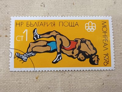 This is a detailed description of a postage stamp featuring a wrestling scene. The stamp has a yellow background with a white serrated border. In the center, two male figures are depicted in a wrestling match. One wrestler is on his back, with his legs in the air, while the other is in a crouched position, grappling with him. Both wrestlers have tan skin and are wearing wrestling attire. The wrestler on his back is wearing blue shorts, and the wrestler in the crouched position is wearing a red singlet.

In the top left corner of the stamp, the Cyrillic text "НР БЪЛГАРИЯ ПОЩА" is visible, which translates to "People's Republic of Bulgaria Post." To the right of this text, there is a white icon of five interlocking Olympic rings. In the bottom left corner, there is a large white numeral "1" with "СТ" below it, indicating a denomination of 1 cent. Along the right edge of the stamp, written vertically in Cyrillic, are the words "МОНРЕАЛ 1976," which translate to "Montreal 1976," suggesting the stamp commemorates the 1976 Montreal Olympics.

The image does not contain any information or visual elements that would indicate a location in the city of Onaldy, Turkmenistan. This stamp is a philatelic item celebrating a sporting event, not a depiction of a geographical location.