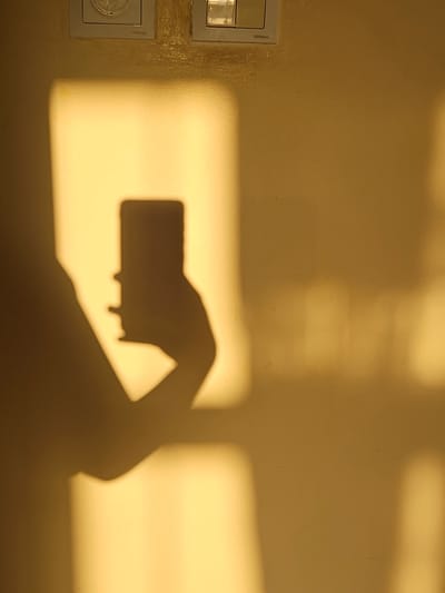 The image depicts a shadow cast on a yellow wall, likely in an indoor setting. The shadow is of a person holding a smartphone, suggesting a selfie is being taken. The light source, creating the shadow, appears to be from a window or artificial light source, casting a warm, golden hue on the wall. Rectangular light patterns are also visible on the wall, indicating the shape of the light source or objects obstructing it. Two electrical outlets or light switches are partially visible at the top of the image, mounted on the wall. The overall scene is simple and focuses on the interplay of light and shadow. The specific location in the city of Saray, Azerbaijan, cannot be determined from the visual information provided in the image.