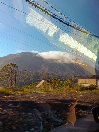 A view from inside a vehicle, looking out at a mountainous landscape in Semangat Gunung, Indonesia. The sky is a clear blue, with several electrical wires crossing the frame diagonally. A reflection on the windshield shows a patterned texture and some green foliage.
In the distance, a large mountain is partially shrouded in white clouds. The slopes of the mountain are covered in dense, dark green forest. In the foreground, to the left, a solitary tree with light green foliage stands on a hillside. Below the mountain, there is a stretch of vegetation, including what appear to be banana plants, and a small building with a textured brown facade.
The ground in the immediate foreground is dark and appears to be dirt or gravel, with some debris scattered across it. The bottom of the image shows the reflection of a person's hand, holding an orange object, likely a pen or stylus, against the interior of the vehicle. The overall impression is a scenic view captured while in transit.