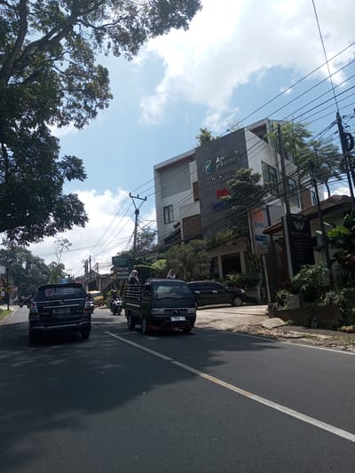 The image depicts a street scene during the daytime. In the foreground, there is a black SUV with license plate "F 1443 KD" and a black pickup truck with license plate "F 8368 YF" driving on the road. There are two women in the back of the pickup truck. Other vehicles and motorcycles are visible further down the road. On the right side of the road is a building complex with a sign that says "Apotek Koshi" and "Klinik Keluarga". The complex also includes an ATM from "BNI". Power lines and trees are seen above and on the sides of the road. The sky is blue with scattered clouds. The road has a white lane marking. The location is likely not Laos.