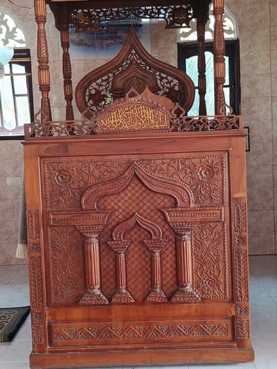 The image depicts a richly carved wooden minbar (a pulpit in a mosque) in the city of Sawang, Indonesia. The minbar is made of dark wood and features intricate Islamic geometric patterns and floral motifs. It has a tall, pointed canopy topped with a finial. The front panel of the minbar is decorated with carved columns and arches, and there is Arabic calligraphy inscribed in gold at the top of the main section. The minbar is positioned in front of a wall covered with light-colored tiles. To the left and right of the minbar, tall windows with decorative grilles are visible. The floor appears to be tiled, and a small portion of a green rug is visible in the lower left corner. The lighting suggests an indoor setting, likely within a mosque.