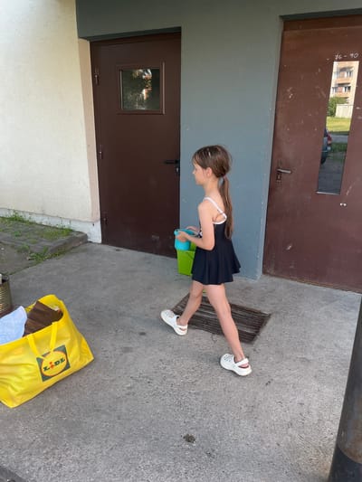 A young girl with a ponytail, wearing a black dress and white Crocs, walks along a concrete pathway. She holds a small blue bucket and a larger green bucket. To her left, a bright yellow Lidl shopping bag is filled with various items, including what appears to be paper and cardboard. The scene is set against a building with two brown doors and a textured grey wall. The environment suggests an outdoor urban setting, possibly in the city of Vangaži, Latvia, given the implied location. The lighting indicates it is daytime.