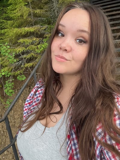 A young woman with long, brown hair is captured in a selfie outdoors. She is wearing a light grey ribbed tank top and a red, white, and blue plaid button-up shirt, unbuttoned and open. Her makeup includes winged eyeliner and subtle eyeshadow. She has freckles across her nose and cheeks. Behind her, a lush green forest with pine trees and some undergrowth is visible, along with a dark metal railing. The background suggests a natural, possibly wooded, setting. There is no indication of the city of Ляды, Russia, in the image.