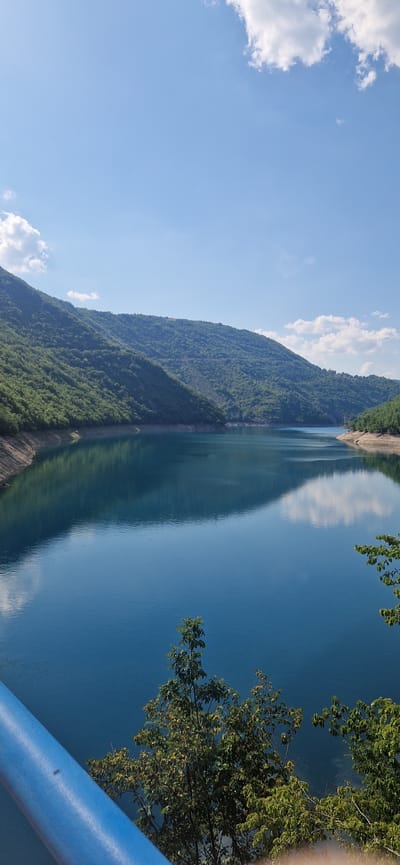 The image shows a panoramic view of a tranquil lake surrounded by verdant, tree-covered hills. The water is a deep blue, reflecting the clear sky and the surrounding landscape. A few white clouds dot the sky, adding to the serene atmosphere. In the foreground, a blue railing is partially visible, suggesting the viewpoint is from a bridge or observation deck. Lush green trees and foliage are also present in the foreground, adding depth to the scene. The overall impression is one of natural beauty and peacefulness. The location is identified as Bezuje, Montenegro, which is known for its stunning natural landscapes and pristine lakes.