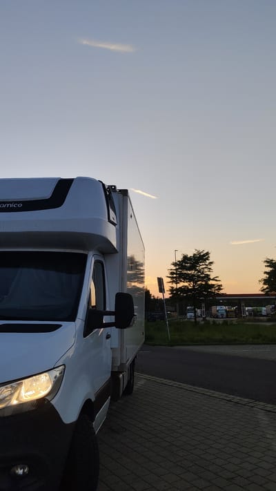 The image depicts a white delivery truck parked on a paved area, possibly a rest stop or parking lot, in the city of Morsleben, Germany. The truck is positioned in the foreground, taking up a significant portion of the left side of the frame. Its headlights are illuminated. In the background, several other trucks are visible parked in front of a building that appears to be a gas station or truck stop. Trees are scattered throughout the background, and the sky shows a soft, gradient of colors characteristic of sunset or sunrise, with a single faint contrail stretching across the upper portion of the sky. The overall scene is calm and still, with no people visible.