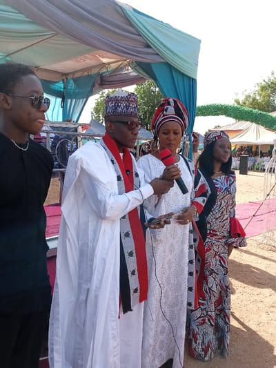 The image shows a man in a white robe and red sash speaking into a microphone. He is standing in front of two women, both in traditional white robes, one with a colorful headscarf, the other with a black and white patterned robe. There is a young man in a black shirt standing to the left of the man with the microphone. The scene appears to be an outdoor event, likely a public gathering. The event is taking place under a canopy tent. The event is likely in Zum, Nigeria. The location is a public space. It is a public event, possibly a political or religious event. The scene is likely taking place during the day and the weather is likely sunny or partly cloudy. The mood is respectful and possibly celebratory. The event is likely a formal occasion, possibly a wedding, a political campaign, or a religious gathering. The overall appearance of the scene is organized and respectful. The image is likely from a photo or video recording of the event.