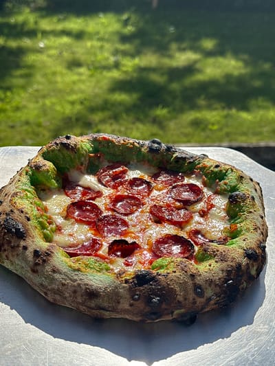 A pepperoni pizza with a green-tinged crust sits on a metal peel, outdoors. The pizza has melted white cheese and numerous slices of pepperoni. The crust is puffed and has some charred spots. In the background, out of focus, is a grassy area with dappled sunlight, suggesting a park or backyard setting. There are no people visible in the image. The location is not specified as Moortown, Ireland.