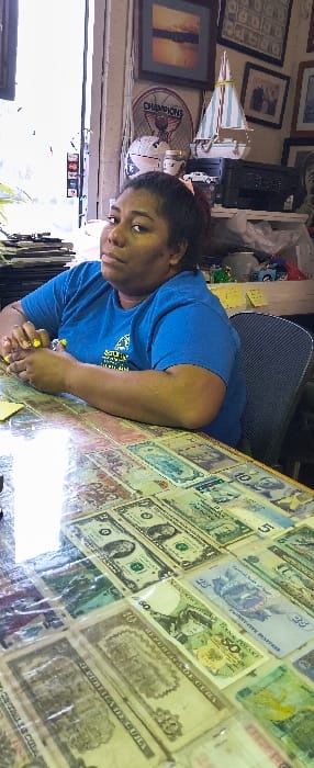 This image depicts an adult female seated at a table within an indoor commercial or office setting in Cutler Bay, United States of America.
The woman, with dark skin and dark hair pulled back, occupies the center-right of the frame, looking directly towards the viewer with a neutral expression. She is seated in a dark chair and wears a short-sleeved blue t-shirt with a yellow logo partially visible on the left chest. Her fingernails are painted yellow, and she has a ring on her right ring finger. Her forearms rest on the table, with her right hand holding a yellow object, possibly a pen or highlighter.
The table surface is covered by a clear protective layer, beneath which numerous international banknotes are displayed. These include US Dollar bills (e.g., $1, $5), a Polish 50 Zloty note, and Cuban 10 Pesos notes, among others. A yellow sticky note is visible on the table near her left hand.
The background, largely behind the woman, features a brightly lit window or doorway on the left, causing some overexposure. To the left of the window, dark stacked items are visible. The wall behind the woman is adorned with various objects: a framed picture depicting a sunset or sunrise scene, a circular "CHAMPIONS" sign with a bee graphic, and a decorative sailboat model with blue and white striped sails. A black printer/scanner unit sits on a surface to the right, surrounded by other miscellaneous items including small toys (e.g., a blue truck), white containers, and plastic bags. Another framed picture containing multiple figures is partially visible to the far right. A yellow sticky note is also present on a surface to the right of the woman's head.